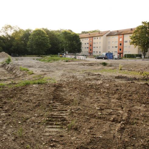 Ein Siedlungsrand mit Bäumen, daneben eine größere Fläche, die bereits als Baustelle vorbereitet ist und auf der die Vegetation entfernt wurde.